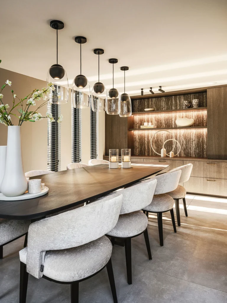 Eettafel van eiken met crèmekleurige stoelen, vijf rookglazen hanglampen en een verlichte kast met bronskleurige wand.