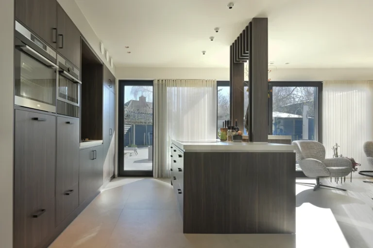 Keuken met donker eiken eiland en licht stenen blad, doorkijk naar de zithoek met bouclé fauteuils bij het raam.