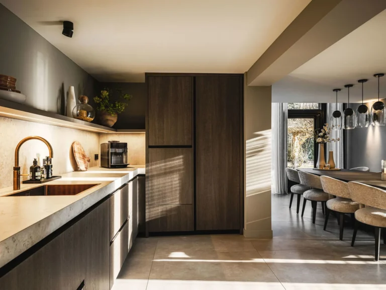 Keuken met licht natuurstenen blad, messing kraan en donker eiken kastenwand, met doorkijk naar de eethoek.