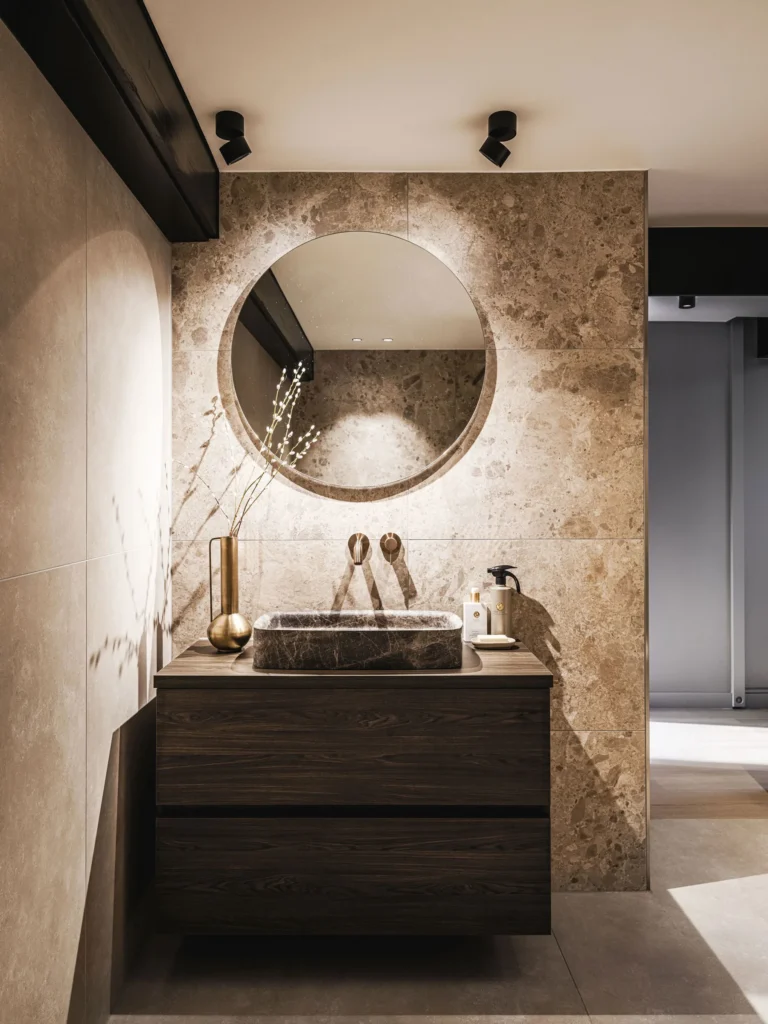 Toilet met wand van travertijn, ronde spiegel met achtergrondlicht, donker houten meubel en marmeren waskom.