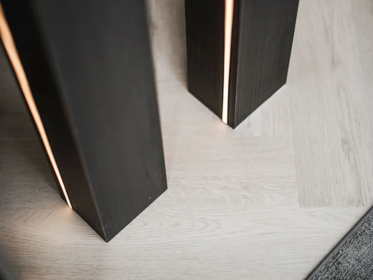 Detail van twee zwart gebeitste eiken staanders met een warme lichtlijn in de naad, op een lichte visgraatvloer.