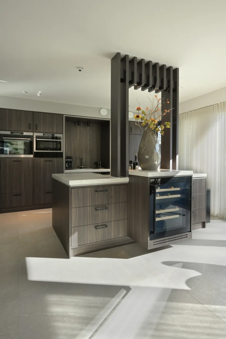 Keuken in donker houtfineer met natuurstenen bladen, ingebouwde wijnklimaatkast en een houten lamellenelement als scheiding.