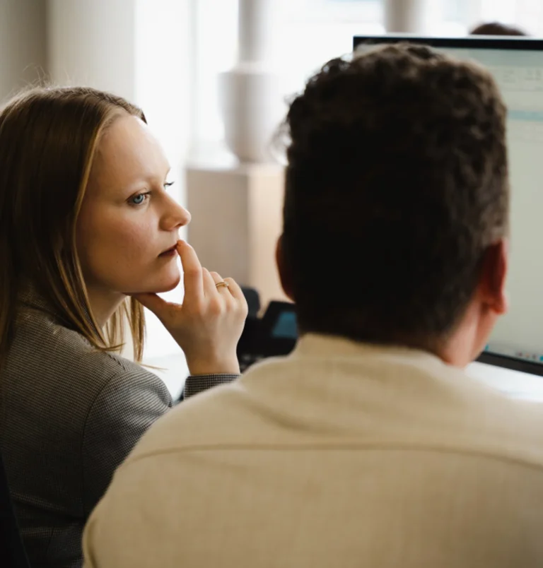 Twee collega's overleggen achter een beeldscherm; een van hen luistert aandachtig met de hand aan haar kin.