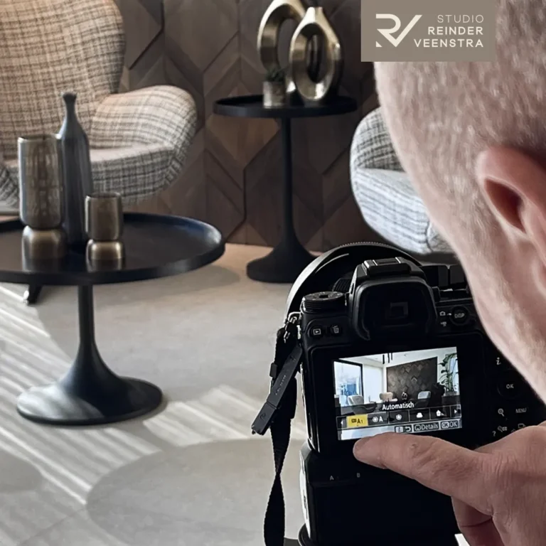 Fotograaf controleert een interieuropname op zijn camera, met achter hem bouclé fauteuils en een visgraat houtwand.